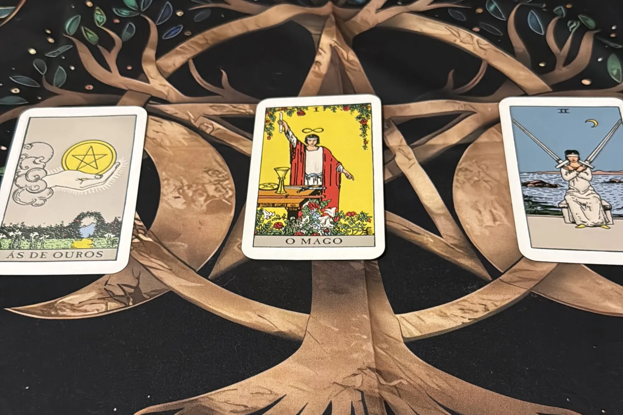 Três cartas de tarot, o Ás de Ouros, o Mago e o Dois de Espadas, sobre um pano com a árvore da vida